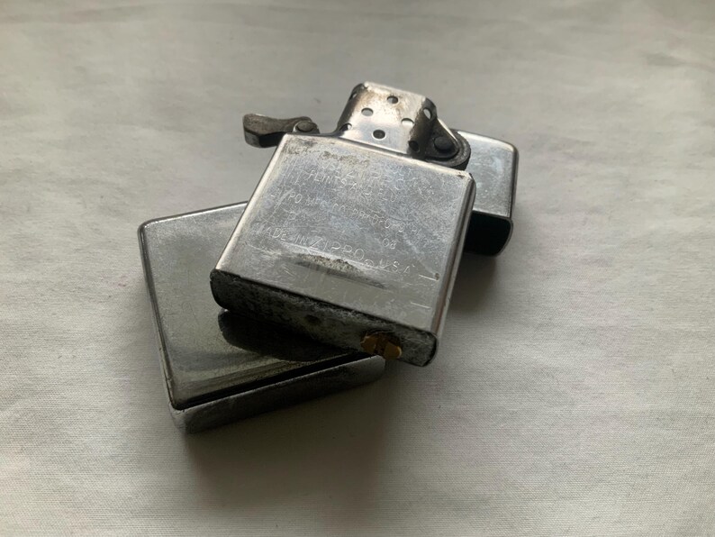 Puede incluir: Dos encendedores Zippo plateados apilados sobre una superficie clara. El encendedor superior est&aacute; abierto, revelando el interior. Los encendedores parecen usados, con algo de desgaste visible en el metal.