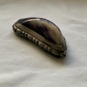 May include: A decorative, antique-style jewelry box. The box has a silver-colored metal frame with ornate detailing around the edges. The top is a smooth, curved shape with a purple and white marbled pattern. The box is approximately 4 inches long.