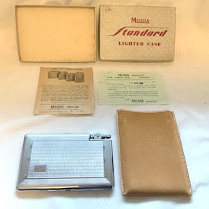 May include: A vintage Mosda Standard lighter case set. The set includes a silver-colored lighter, a tan leather pouch, the original box, and service documents. The box is beige with red text. The lighter has a textured design.