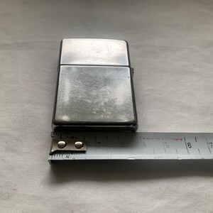 Puede incluir: Un encendedor de metal plateado con forma rectangular. El encendedor est&aacute; colocado junto a una cinta m&eacute;trica, que muestra una longitud de aproximadamente 5,7 cm.