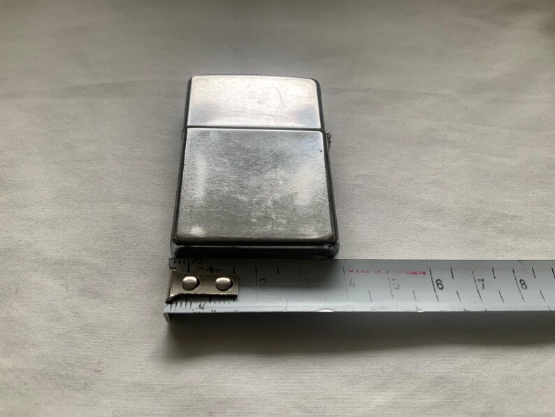 Puede incluir: Un encendedor de metal plateado con forma rectangular. El encendedor est&aacute; colocado junto a una cinta m&eacute;trica, que muestra una longitud de aproximadamente 5,7 cm.