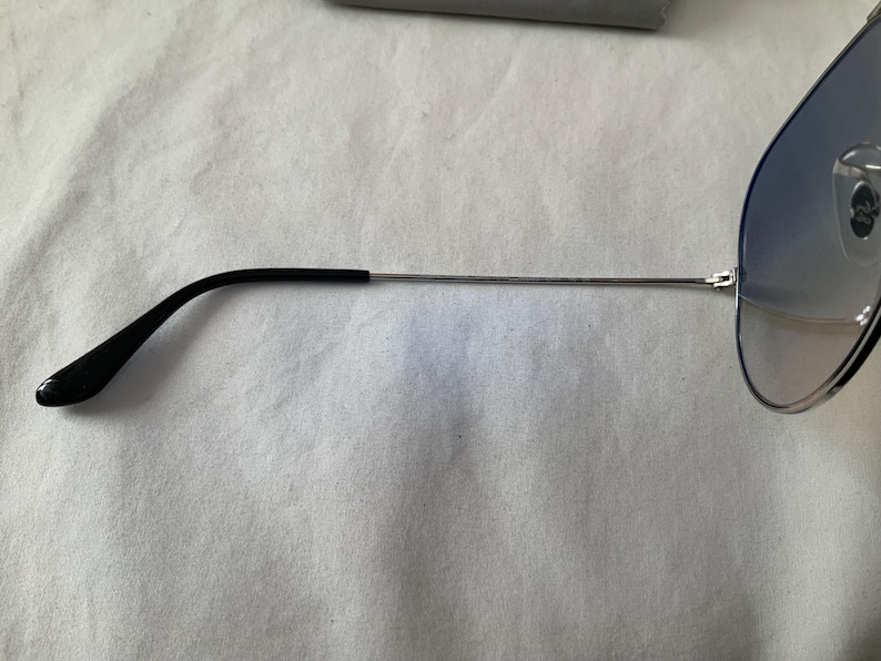 May include: Aviator-style sunglasses with a silver frame and gradient blue lenses. The temples are black and extend to the ear. The sunglasses are partially visible against a white background.