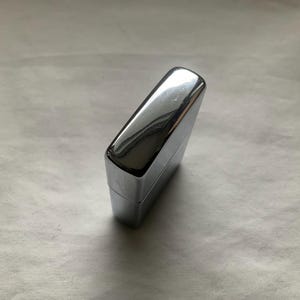 May include: A shiny, chrome-colored lighter with a rectangular shape. The lighter has a smooth, reflective surface, and the top is slightly rounded. The lighter is closed and appears to be new.