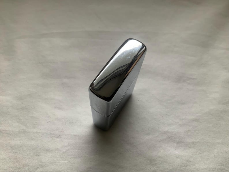 May include: A shiny, chrome-colored lighter with a rectangular shape. The lighter has a smooth, reflective surface, and the top is slightly rounded. The lighter is closed and appears to be new.