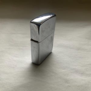 May include: A shiny, silver-colored metal lighter. The rectangular lighter has a hinged lid and a smooth, reflective surface. The lighter is closed and stands upright on a neutral-colored surface.