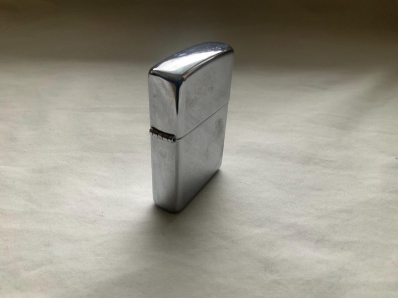May include: A shiny, silver-colored metal lighter. The rectangular lighter has a hinged lid and a smooth, reflective surface. The lighter is closed and stands upright on a neutral-colored surface.