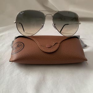 May include: Silver-framed aviator sunglasses with gradient lenses rest on a brown leather case. The case has a snap closure and a debossed logo. The sunglasses have a classic teardrop shape and a thin metal frame. The lenses transition from dark to light.