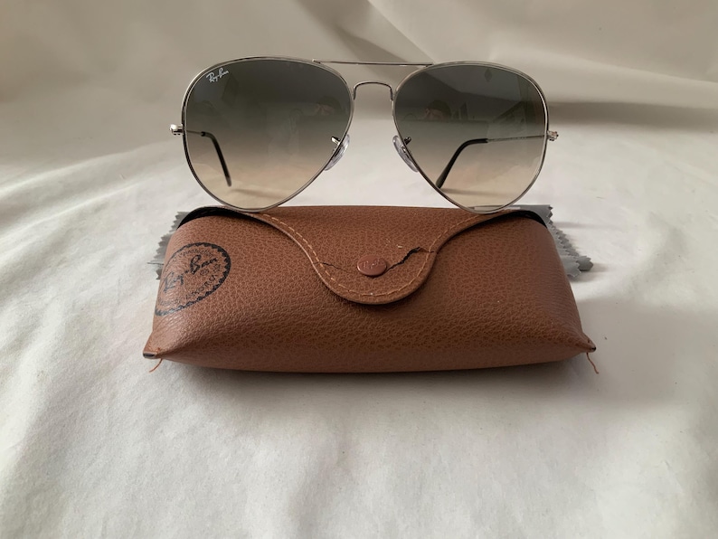 May include: Silver-framed aviator sunglasses with gradient lenses rest on a brown leather case. The case has a snap closure and a debossed logo. The sunglasses have a classic teardrop shape and a thin metal frame. The lenses transition from dark to light.