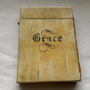 Könnte beinhalten: Eine kleine, rechteckige, elfenbeinfarbene Schachtel mit einem Scharnierdeckel. Die Schachtel trägt eine schwarze Inschrift mit der Aufschrift "Grace" in einer dekorativen Schriftart.