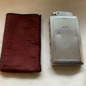 May include: A silver cigarette lighter with a black felt case. The lighter has a textured surface and the initials "GF" engraved on the front.