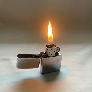 May include: A silver metal lighter with a flame. The lighter is open, revealing the interior. The flame is orange and yellow, and the lighter has a small, patterned top.