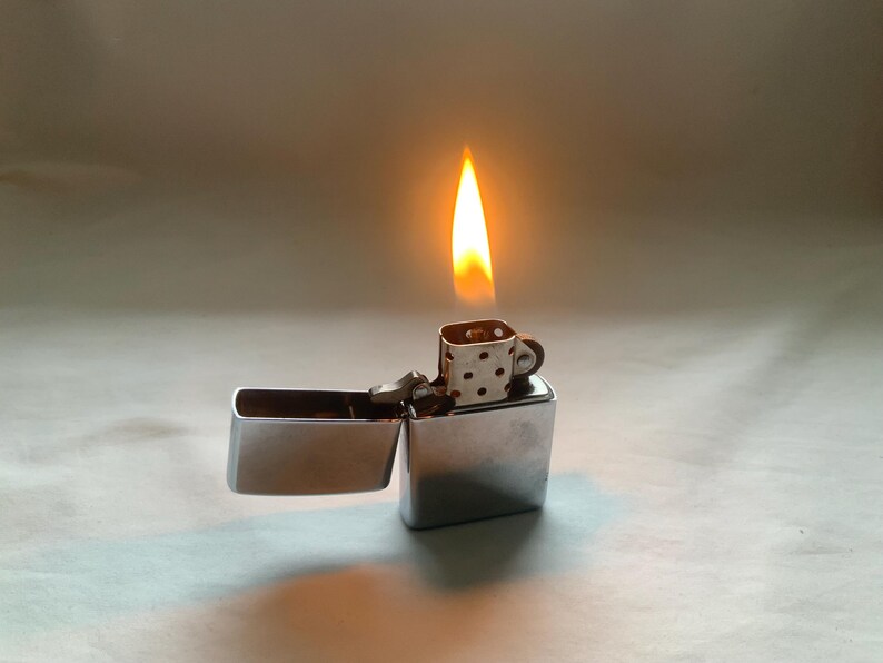 May include: A silver metal lighter with a flame. The lighter is open, revealing the interior. The flame is orange and yellow, and the lighter has a small, patterned top.