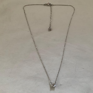 Peut inclure: Un collier argenté avec une chaîne délicate et un seul pendentif en cristal clair de forme carrée. Le collier a une longueur réglable et un petit charme décoratif près du fermoir. Le bijou est présenté sur une surface claire.