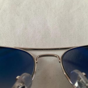 May include: Aviator-style sunglasses with a silver frame and gradient blue lenses. The top bar of the frame has the text "MADE IN FRANCE". The lenses are a deep blue color, and the frame has a classic, sleek design.