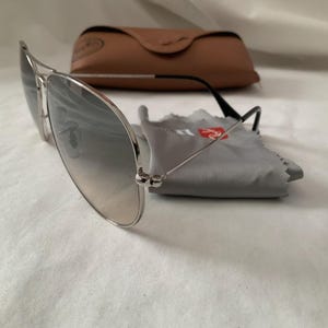 May include: Silver-framed aviator sunglasses with gradient lenses, a brown leather case, and a gray cleaning cloth. The sunglasses have black temples and are resting on a white surface. The case is closed.
