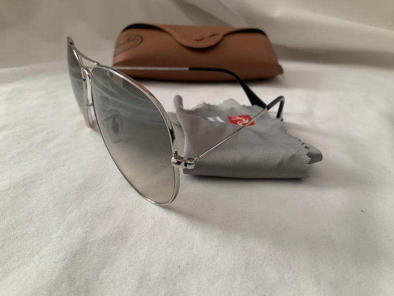 May include: Silver-framed aviator sunglasses with gradient lenses, a brown leather case, and a gray cleaning cloth. The sunglasses have black temples and are resting on a white surface. The case is closed.