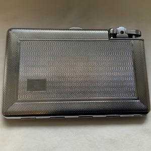May include: A rectangular, silver-toned cigarette case with a built-in lighter. The case has a textured, patterned surface with a small rectangular accent. The lighter is located on the top right corner.