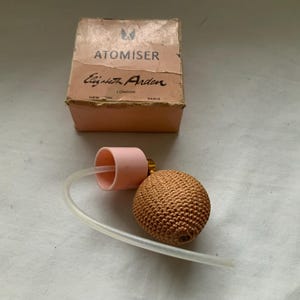 Puede incluir: Un frasco atomizador rosa y marrón con el texto "ATOMISER Elizabeth Arden NEW YORK LONDON PARIS" en la caja.