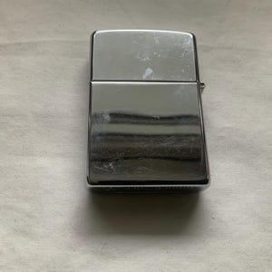 May include: A rectangular, chrome-plated lighter with a hinged lid. The lighter has a shiny, reflective surface with visible scratches. The design is simple and classic, with a small hinge on the side.