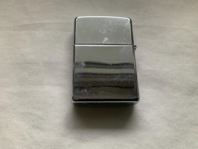 May include: A rectangular, chrome-plated lighter with a hinged lid. The lighter has a shiny, reflective surface with visible scratches. The design is simple and classic, with a small hinge on the side.
