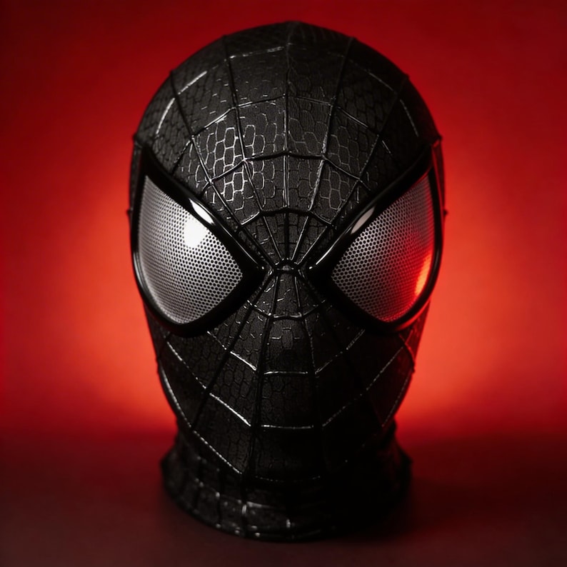May include: A black Spider-Man mask with a textured surface and web detailing. The mask features large, white mesh eye lenses and is set against a red background. The mask is a replica of the superhero's costume.