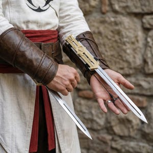 K&ouml;nnte beinhalten: Zwei silberne Klingen erstrecken sich von einem gold-schwarzen Mechanismus, der an einer braunen Lederarmschiene befestigt ist. Die Armschienen werden am Unterarm getragen, mit einem roten Sch&auml;rpenband und wei&szlig;er Tunika im Hintergrund.