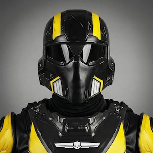 Puede incluir: Casco futurista negro y amarillo con visera. El casco tiene rayas amarillas en la parte superior y detalles amarillos en los laterales. El diseño incluye una máscara facial negra y un emblema de calavera en el pecho.