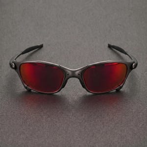 Può includere: Occhiali da sole grigio scuro con lenti rosse. Gli occhiali hanno un design elegante e moderno con una montatura e aste curve. Le lenti sono di un colore rosso vibrante, offrendo un audace contrasto con la montatura.