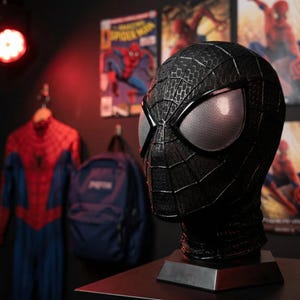 May include: A black Spider-Man mask with a textured surface and silver mesh eye lenses. The mask is displayed on a dark base. In the background, a Spider-Man costume hangs on a rack, along with a backpack and comic book posters.