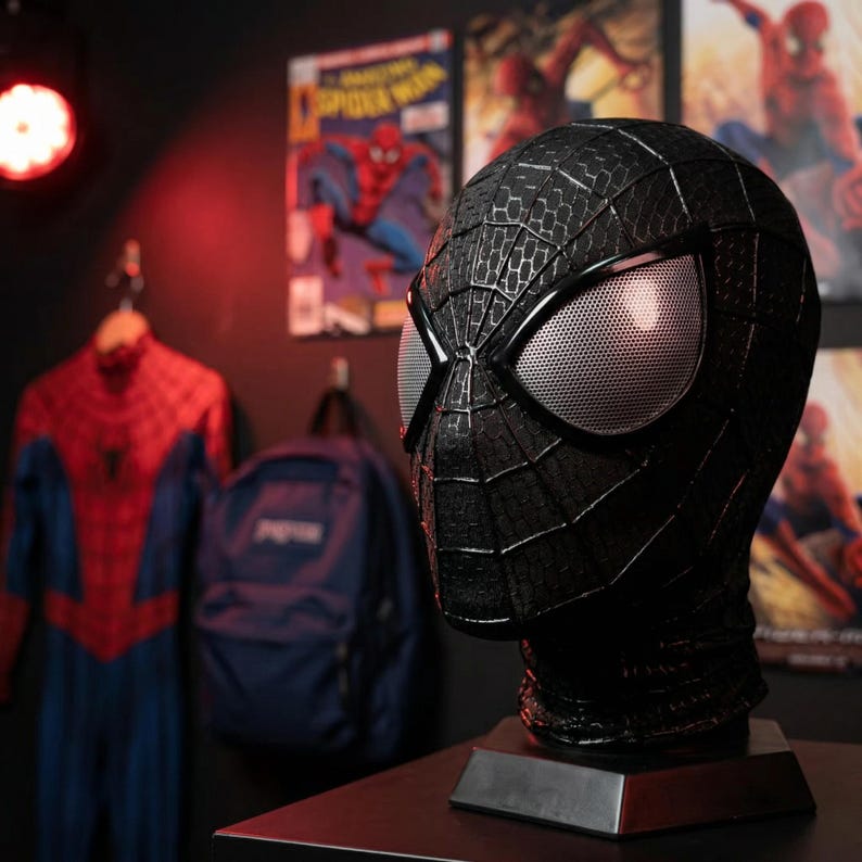 May include: A black Spider-Man mask with a textured surface and silver mesh eye lenses. The mask is displayed on a dark base. In the background, a Spider-Man costume hangs on a rack, along with a backpack and comic book posters.