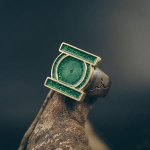 Puede incluir: Un anillo con un marco rectangular dorado y un centro circular, relleno de esmalte verde texturizado y un patrón de espiral central. La banda es de metal oscuro y desgastado con una textura similar a una grieta en la superficie lateral.