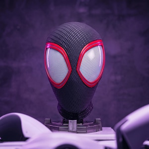 Könnte beinhalten: Eine schwarze Miles Morales Spider-Man Maske mit einer texturierten Oberfläche mit wiederkehrendem Muster, großen weißen Netz-Augenlinsen und leuchtend roten Umrandungen um die Augenöffnungen, montiert auf einem dunkelgrauen Präsentationsständer.