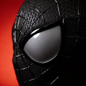 May include: Close-up of a black Spider-Man mask with white mesh eye lenses. The mask has a textured, web-like pattern and is set against a red background. The eye lenses are framed by a glossy black material.