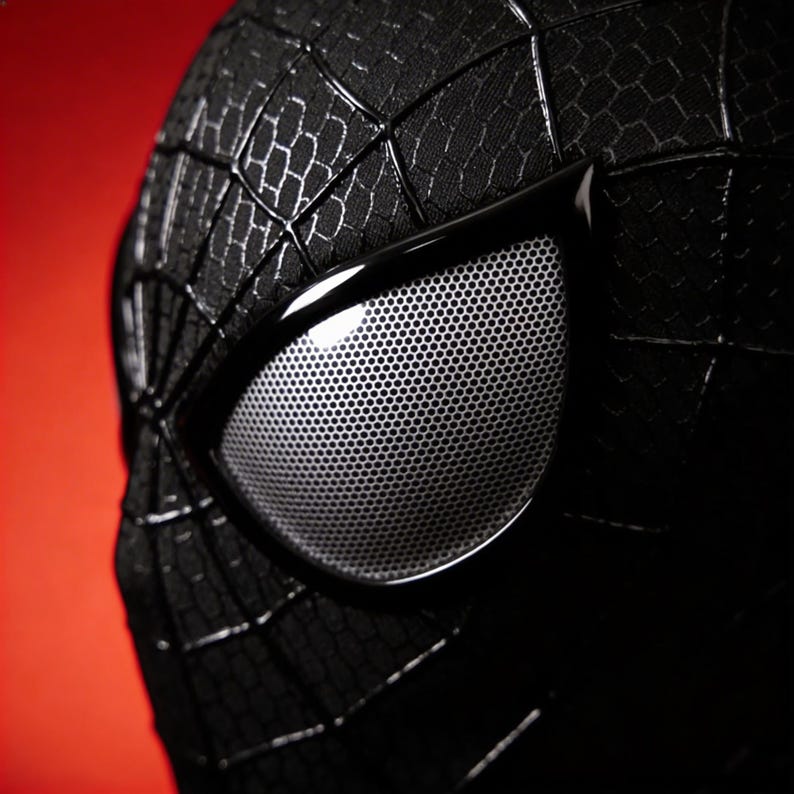 May include: Close-up of a black Spider-Man mask with white mesh eye lenses. The mask has a textured, web-like pattern and is set against a red background. The eye lenses are framed by a glossy black material.