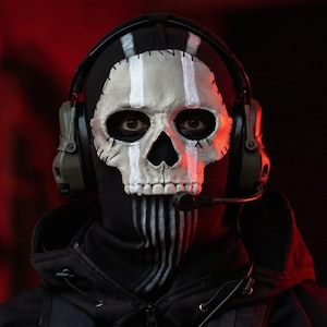 May include: A black hooded mask with a white skull design, vertical white stripes, and a headset with a microphone. The mask covers the face, with the skull design prominently displayed. The background is dark red.