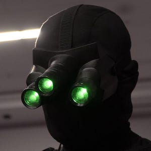 Puede incluir: Un atuendo táctico negro con gafas de visión nocturna con tres lentes verdes brillantes. El atuendo incluye una capucha negra, una cobertura facial y una chaqueta con cremallera y bolsillos. La imagen está en escala de grises.