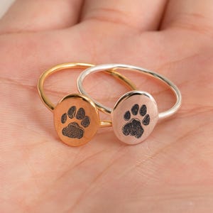 Puede incluir: Dos anillos con diseños de huellas de pata. Uno es dorado y el otro plateado. Cada anillo presenta un pequeño dije ovalado con una huella de pata negra. Anillos sencillos y elegantes, adecuados para el uso diario.