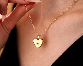May include: A gold-colored heart-shaped locket necklace with a delicate chain. The locket features a small blue gemstone and a starburst design. The necklace is worn against a dark background.