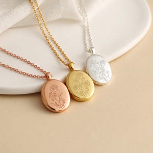 May include: Three oval locket necklaces in rose gold, yellow gold, and silver tones. Each locket features a delicate, fine-line floral engraving on the front surface and hangs from a matching thin metal chain with small, uniform circular links.