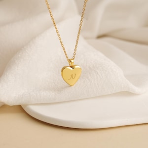 K&ouml;nnte beinhalten: Eine goldfarbene herzf&ouml;rmige Medaillon-Halskette mit feiner Kette. Das Medaillon hat den Buchstaben "N" auf der Vorderseite eingraviert. Die Halskette wird auf einer wei&szlig;en Oberfl&auml;che pr&auml;sentiert.