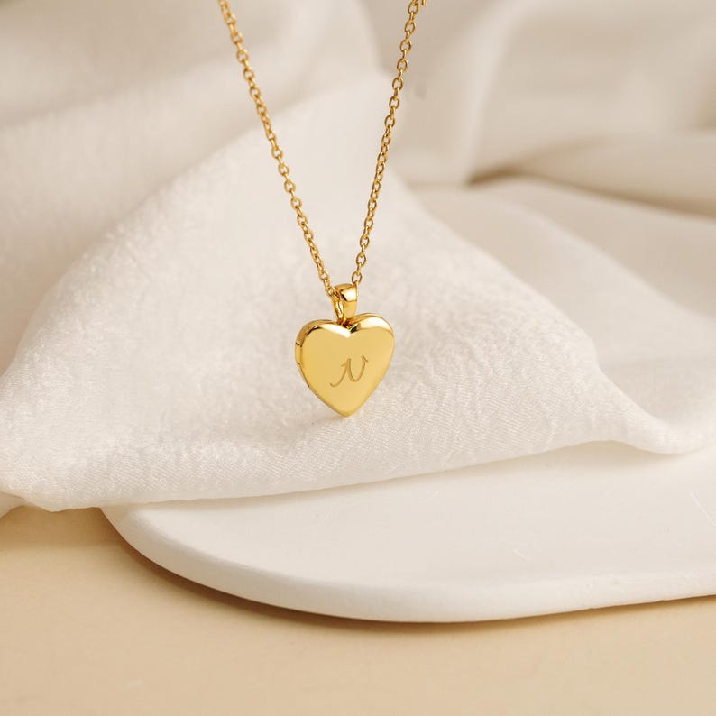 K&ouml;nnte beinhalten: Eine goldfarbene herzf&ouml;rmige Medaillon-Halskette mit feiner Kette. Das Medaillon hat den Buchstaben "N" auf der Vorderseite eingraviert. Die Halskette wird auf einer wei&szlig;en Oberfl&auml;che pr&auml;sentiert.