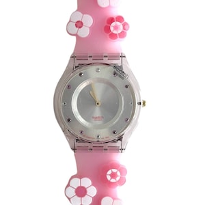 Puede incluir: Un reloj Swatch rosa con una esfera plateada y una caja de plástico transparente. El reloj tiene una correa de silicona rosa con detalles de flores blancas y rosas.