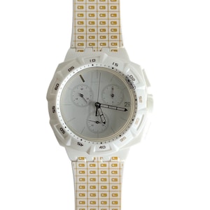 Op de afbeelding: Witte polshorloge met een witte wijzerplaat en goudkleurige wijzers. Het horloge heeft een witte band met een goud-wit geruit patroon. De wijzerplaat van het horloge heeft een chronograaffunctie en de tekst "Swatch" is op de wijzerplaat gedrukt.