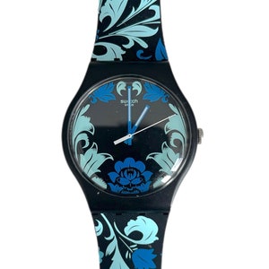 Pode incluir: Um relógio Swatch preto com um padrão floral azul e branco na pulseira. O mostrador do relógio é preto com ponteiros azuis e um logotipo Swatch branco.