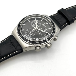 May include: A stainless steel Swatch chronograph watch with a black dial and subdials. The watch features a black leather band with white stitching. The watch has silver-colored buttons and a bezel with white markings.