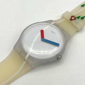May include: A Swatch watch with a transparent gray case and a light-colored band. The watch face is silver with red and blue hands. The band has a pattern of white stripes, green trees, and a brown bear.