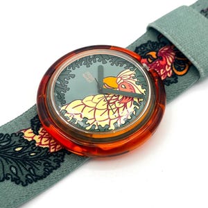 Op de afbeelding: Een Swatch-horloge met een ronde, oranje getinte kast en een lichtgroene wijzerplaat met een geel en oranje vogelontwerp. Het horloge heeft een lichtgroene stoffen band met een herhalend bloemmotief in rood en oranje.