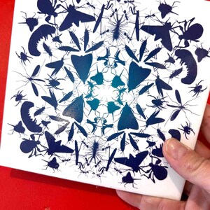 May include: A white ceramic tile with a blue and teal mandala design featuring various insect silhouettes. The design is symmetrical and features a variety of insects, including butterflies, moths, beetles, and ants.