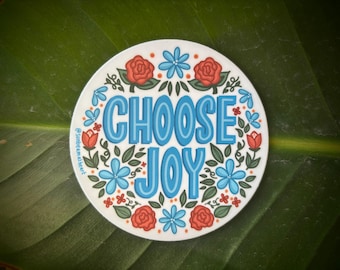 May include: A circular sticker featuring the words "CHOOSE JOY" in blue, encircled by red roses, blue flowers, and green foliage. The sticker is white and set against a green leaf.
