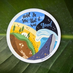 May include: A round sticker with a colorful illustration. The design features a whale, land, and sky with the text "and He saw that it was Good". The sticker has a white border and is set against a green leaf.